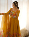 Woman wearing a yellow floral dress with a matching dupatta, standing against a white curtain background.