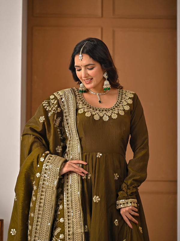Woman wearing a traditional green outfit with intricate embroidery in front of a brown wall.