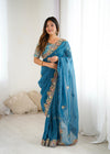 Woman wearing a blue saree with intricate designs in a white room.